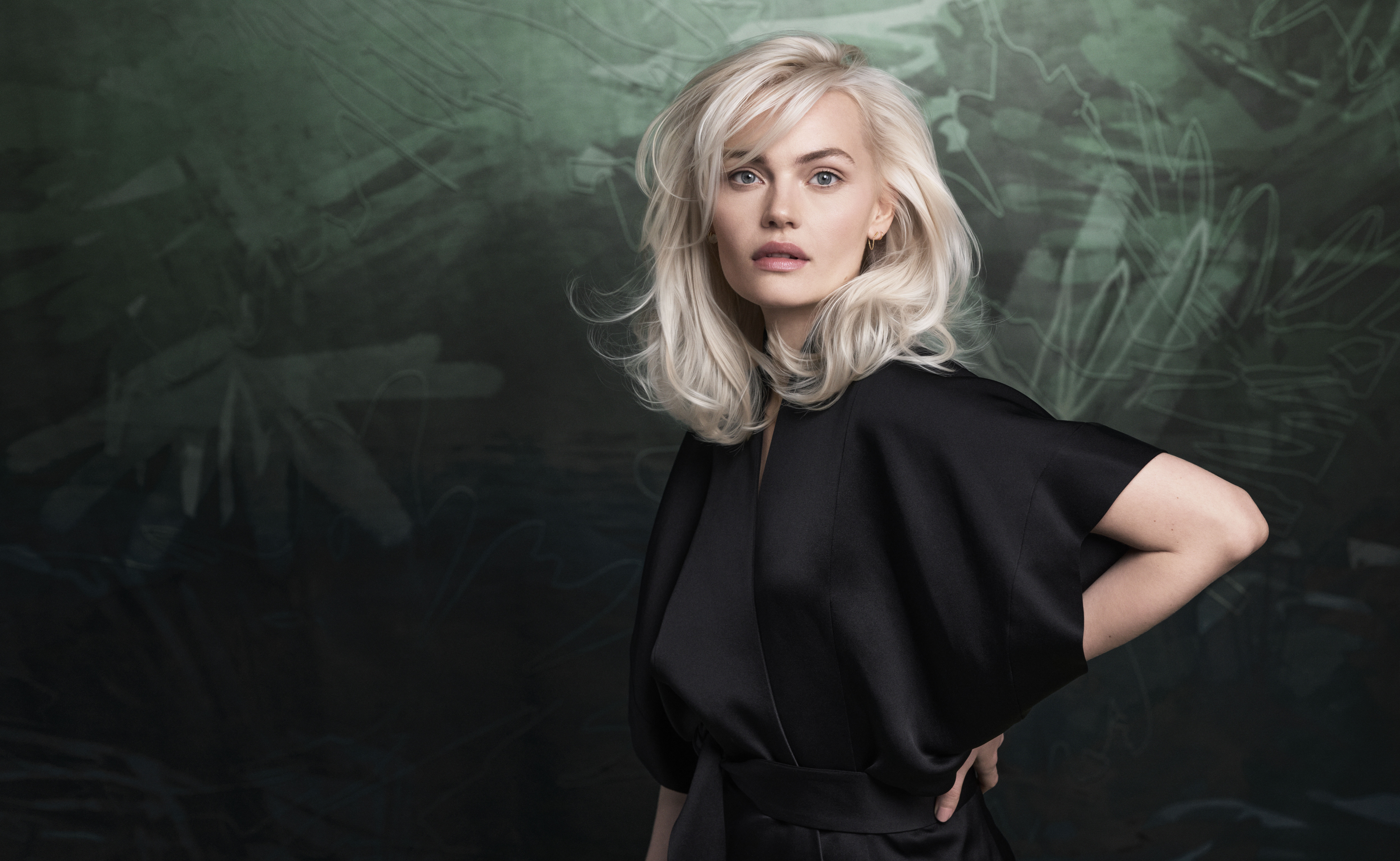 A woman with platinum blonde hair poses against a dark green and black abstract background, wearing a loose-fitting black outfit and holding Aveda pink ribbon hand cream while looking directly at the camera. - Trios Aveda Salon | Fort Collins, CO