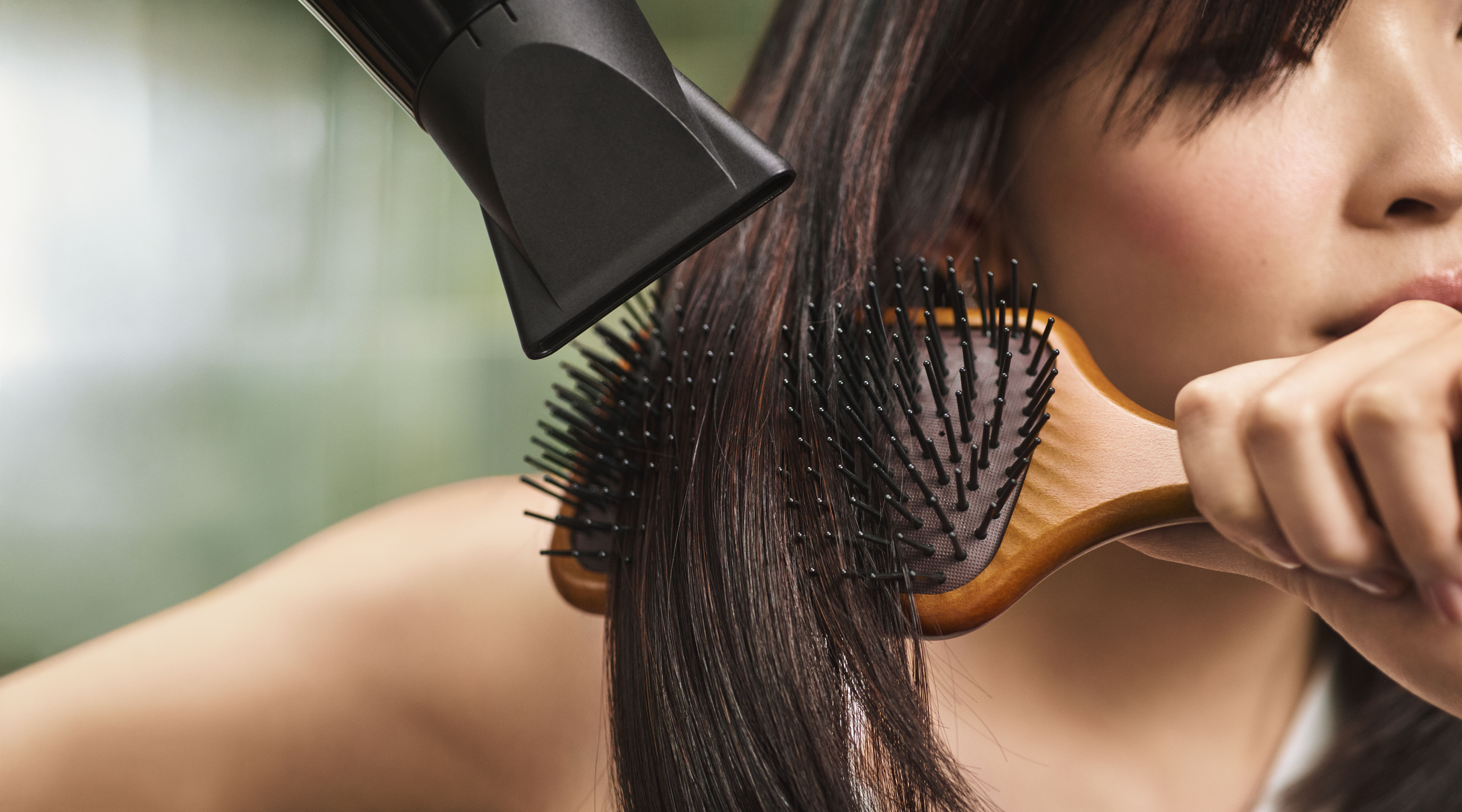 A person dries and brushes their straight dark hair using a hairdryer and a wooden paddle brush, then nourishes their hands with Aveda breast cancer hand relief for soft, smooth skin. - Trios Aveda Salon | Fort Collins, CO