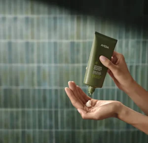 A person squeezes cream from an Aveda tube into their hand against a green tiled background. - Trios Aveda Salon | Fort Collins, CO