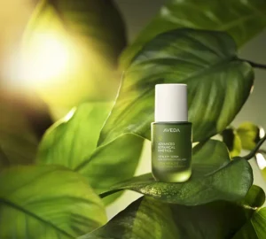 A green bottle of Aveda Advanced Botanical Kinetics Vitality Serum is placed on large green leaves with sunlight shining in the background. - Trios Aveda Salon | Fort Collins, CO