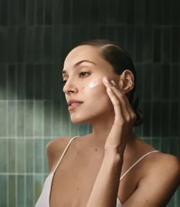 A woman applies a dab of Advanced Botanical Kinetics™ cream to her cheek with her fingers, standing in front of a green tiled wall—perfect for spring hair and skin prep with Aveda skincare. - Trios Aveda Salon | Fort Collins, CO