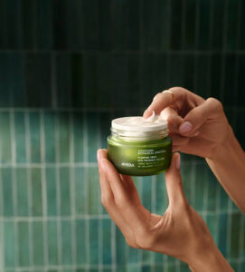 A person holds an open jar of Aveda Botanical Repair Intensive Strengthening Masque—an essential in any Aveda skincare routine—against a green tiled background. - Trios Aveda Salon | Fort Collins, CO