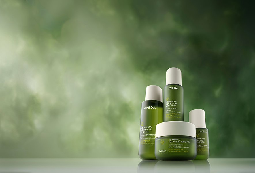 Four green Aveda skin care products are arranged on a reflective surface against a blurred green and white background. - Trios Aveda Salon | Fort Collins, CO