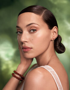 Woman with slicked-back dark hair in a low bun, wearing a white top, small hoop earring, and red beaded bracelet, posing with hand on face against a green blurred background—reflecting the glow of Aveda facials Fort Collins. - Trios Aveda Salon | Fort Collins, CO