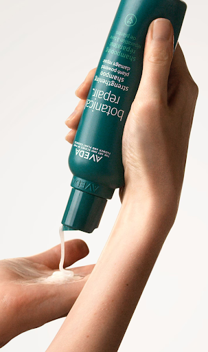 A person is squeezing a green bottle of botanical repair shampoo into their hand against a plain white background. - Trios Aveda Salon | Fort Collins, CO