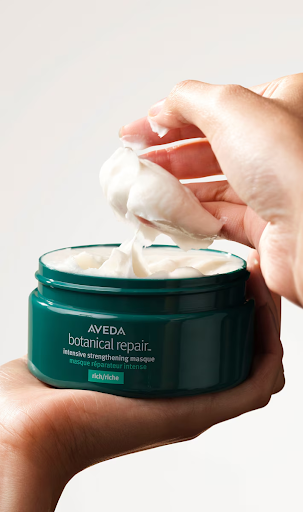 A person's hand scooping cream from an Aveda botanical repair intensive strengthening masque container. - Trios Aveda Salon | Fort Collins, CO