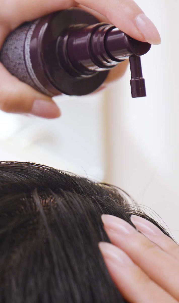 A hand applies a hair care product from a pump dispenser to a person's scalp. - Trios Aveda Salon | Fort Collins, CO