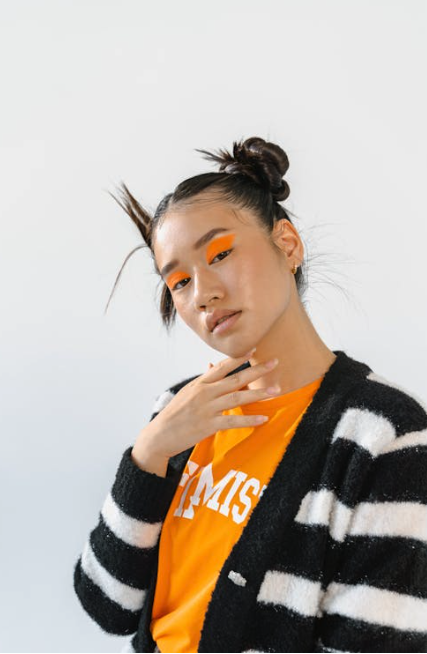 Person with a striped sweater and orange shirt poses with fingers near chin. Hair styled in double buns, with bold orange eyeshadow. Neutral background. - Trios Aveda Salon | Fort Collins, CO