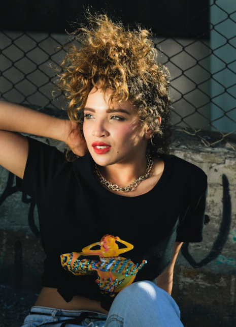 A person with curly hair and red lipstick leans against a graffiti-covered wall, wearing a black t-shirt with a colorful graphic and a gold chain necklace. - Trios Aveda Salon | Fort Collins, CO