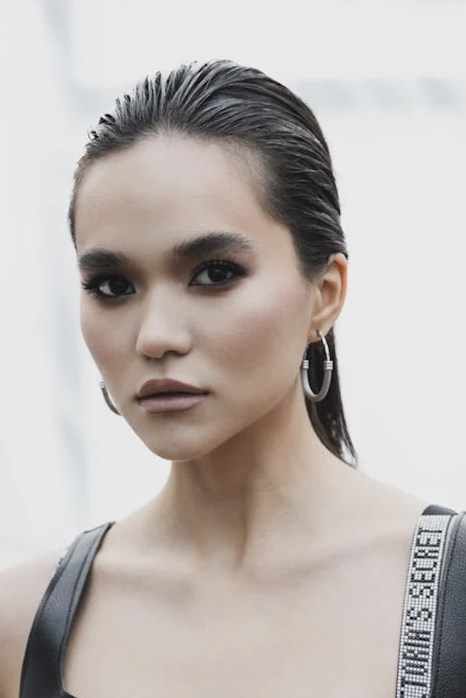 Person with slicked-back hair wearing hoop earrings and a black top, facing forward with a neutral expression. - Trios Aveda Salon | Fort Collins, CO