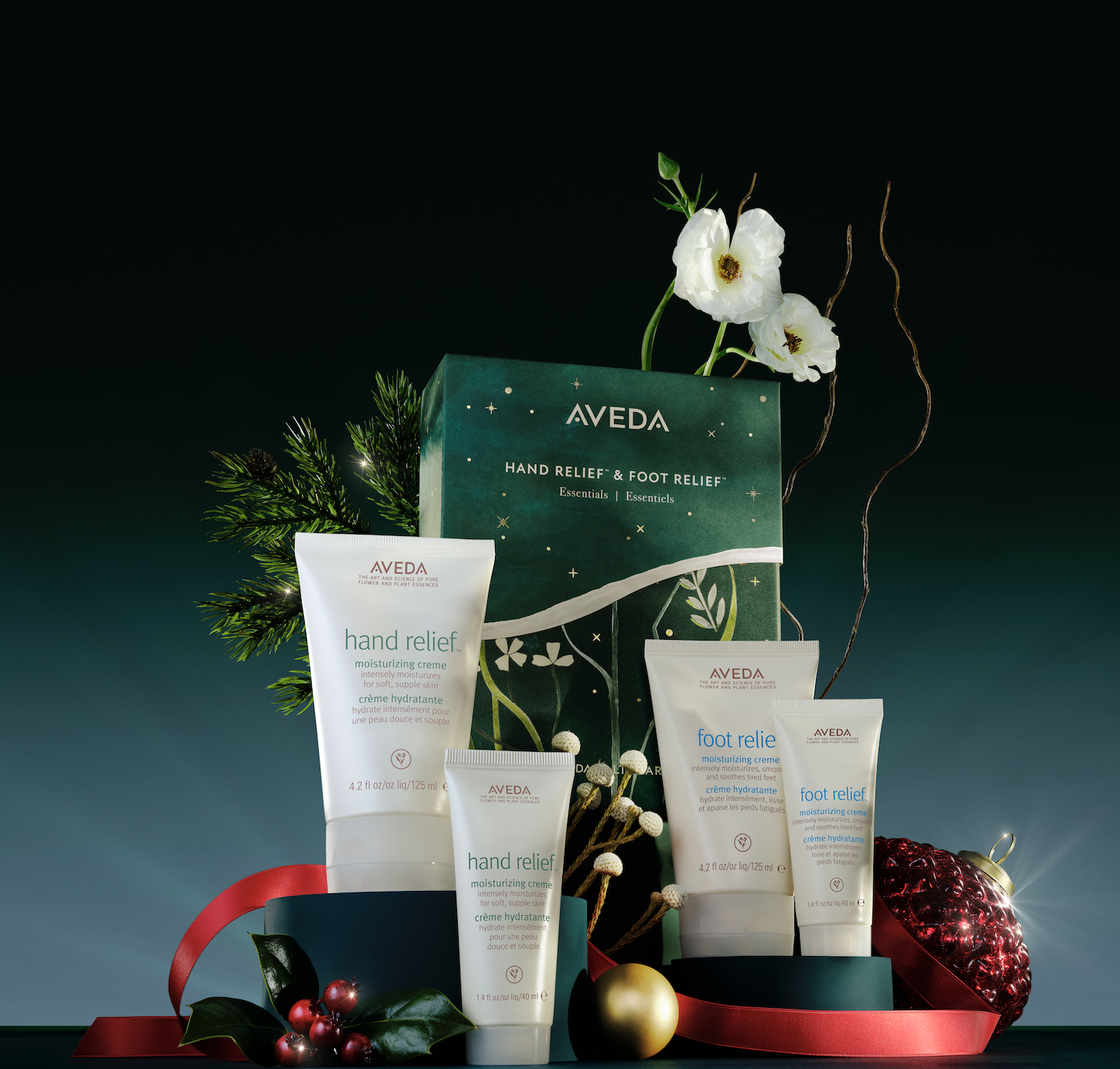 Aveda gift set with hand and foot relief creams, decorative greenery, red ribbon, and ornaments on a dark background. - Trios Aveda Salon | Fort Collins, CO