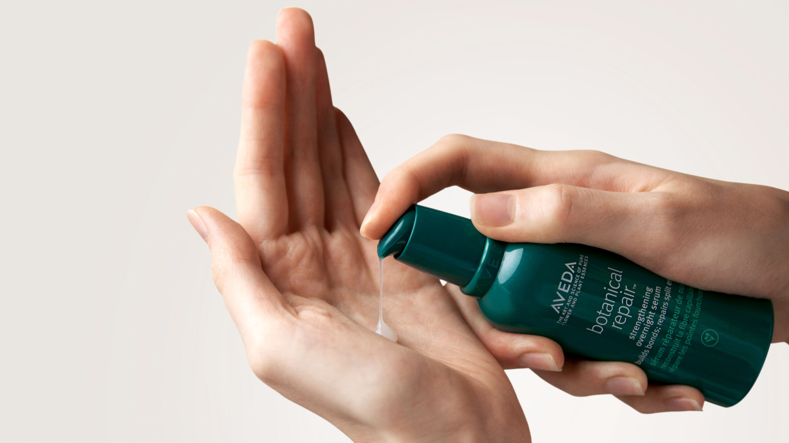 A person dispenses Aveda botanical repair serum into their palm from a green bottle against a plain background. - Trios Aveda Salon | Fort Collins, CO