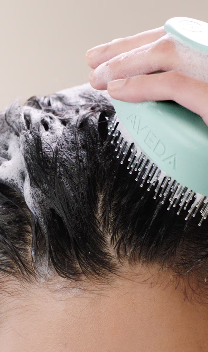 A person uses a scalp massager to apply shampoo to their wet hair, creating suds. - Trios Aveda Salon | Fort Collins, CO