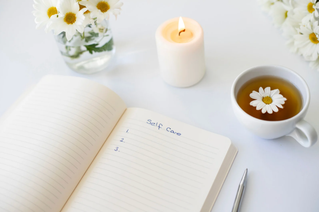 Open notebook with "Self Care" list, a lit candle, daisy-filled cup of tea, and flowers in a vase on a white surface. - Trios Aveda Salon | Fort Collins, CO