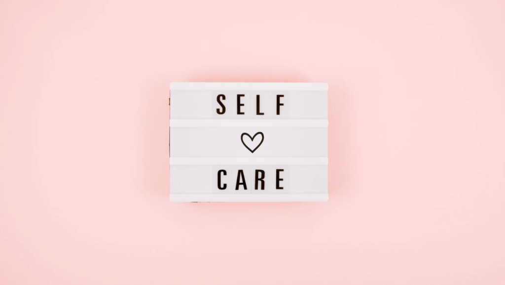 A lightbox against a pink background displays the words "SELF CARE" with a heart symbol between the words. - Trios Aveda Salon | Fort Collins, CO