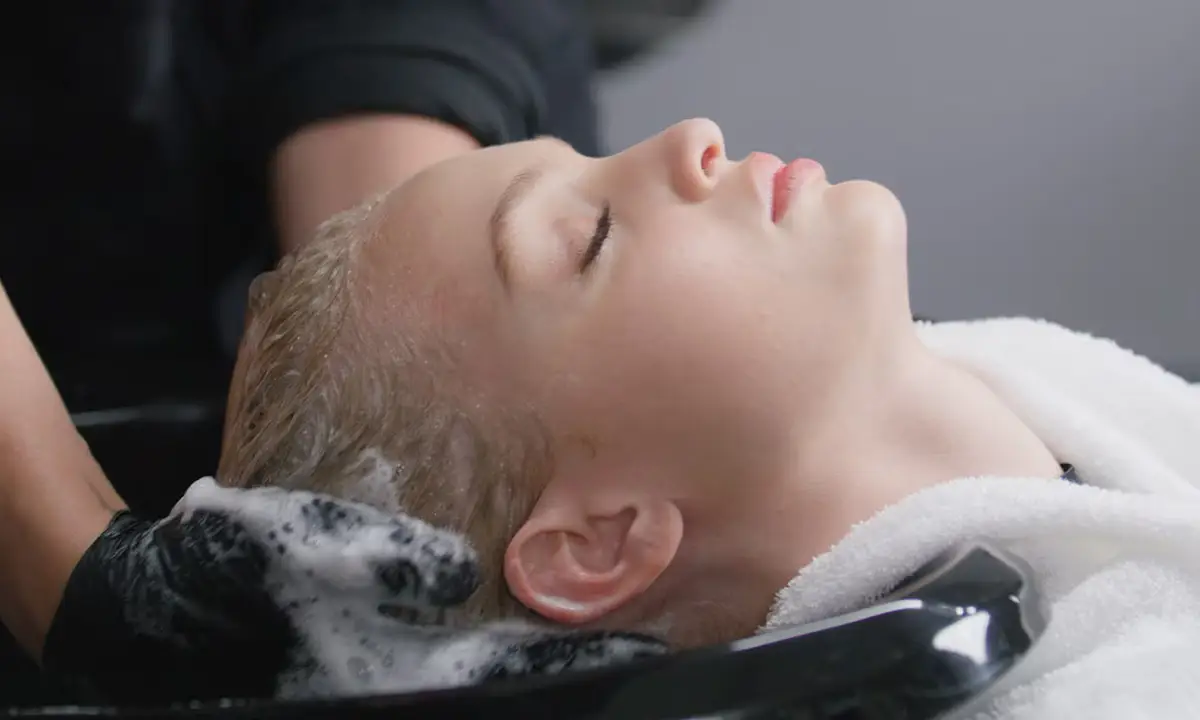 A person with closed eyes reclines at a salon sink while their hair is being washed and lathered with shampoo by someone wearing black gloves. - Trios Aveda Salon | Fort Collins, CO