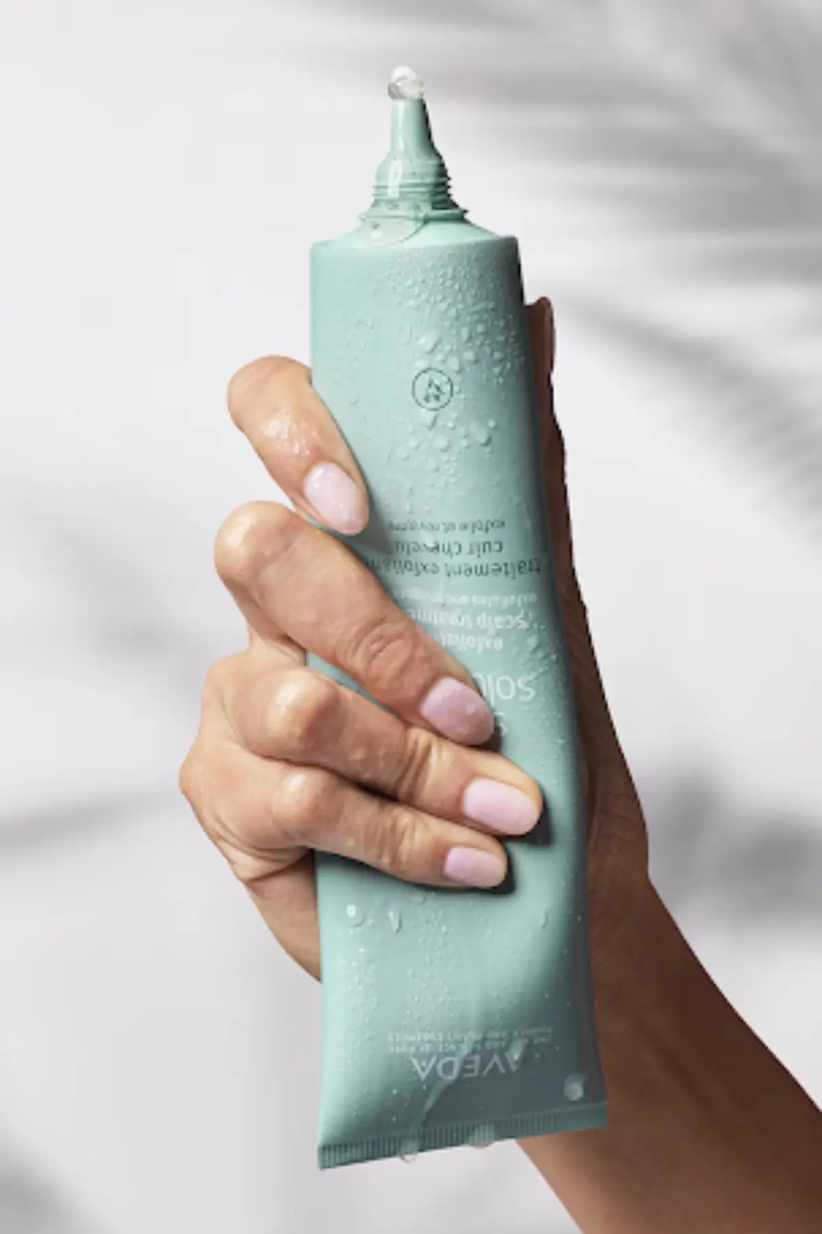 A hand with light pink nails holds a teal tube with some water droplets, squeezing out a small amount of product from the nozzle. - Trios Aveda Salon | Fort Collins, CO