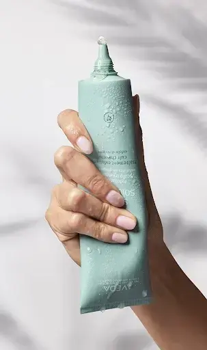 A hand holds a light blue-green tube of Aveda hair care product, with a drop of product visible at the open tip—perfect for spring self-care and nourishing scalp-care routines. - Trios Aveda Salon | Fort Collins, CO