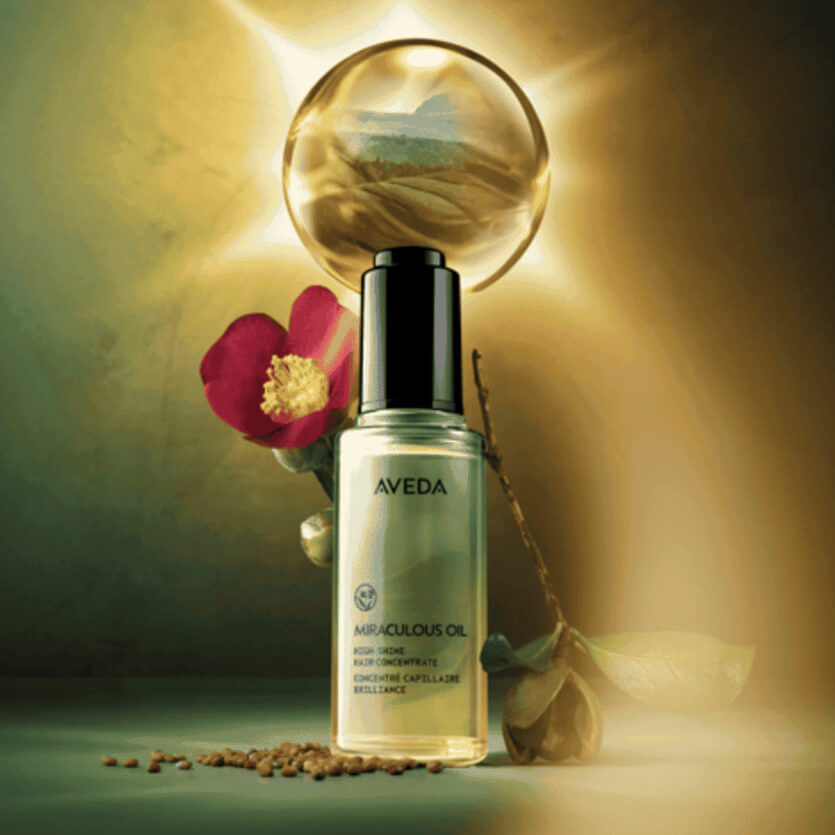 A bottle of Aveda Miraculous Oil is displayed with a flower, seeds, and a glowing orb in the background. - Trios Aveda Salon | Fort Collins, CO
