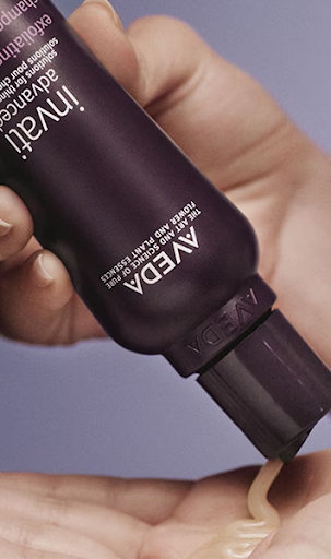A person squeezes a creamy product from a dark purple Aveda tube into their hand. - Trios Aveda Salon | Fort Collins, CO