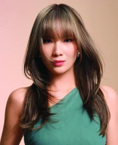 A woman with long, layered brown hair and bangs, wearing a sleeveless green top, poses against a neutral background. - Trios Aveda Salon | Fort Collins, CO