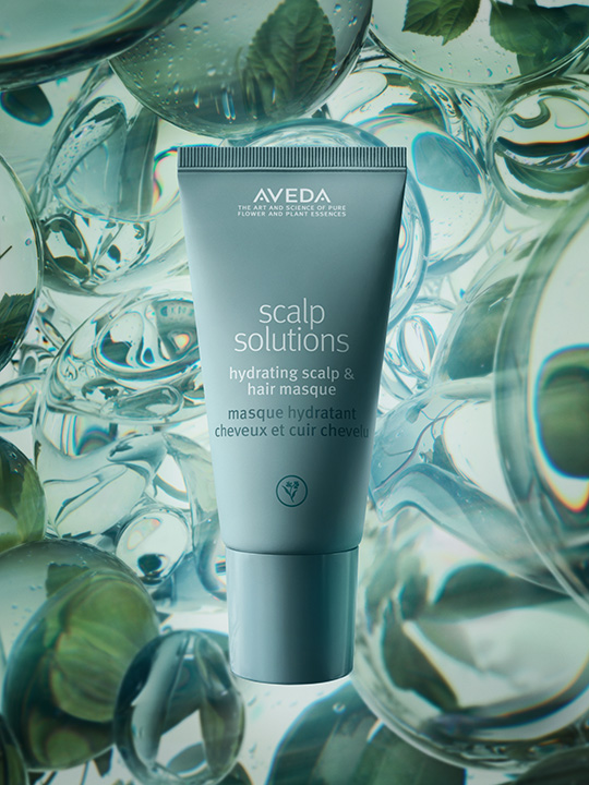 A tube of Aveda Scalp Solutions Hydrating Scalp & Hair Masque stands upright against a background of water droplets and green leaves. - Trios Aveda Salon | Fort Collins, CO