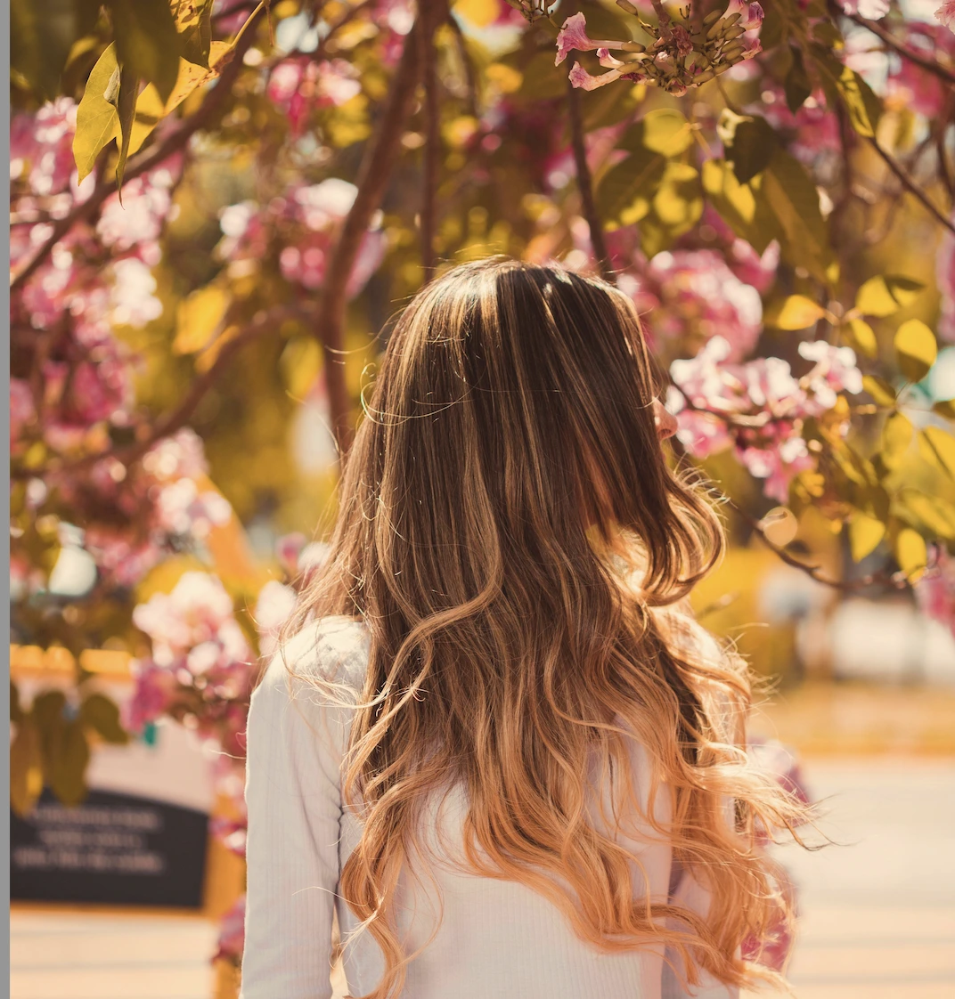 A person with long, wavy, blonde-brown hair stands outdoors facing blooming pink flowers on a tree. - Trios Aveda Salon | Fort Collins, CO