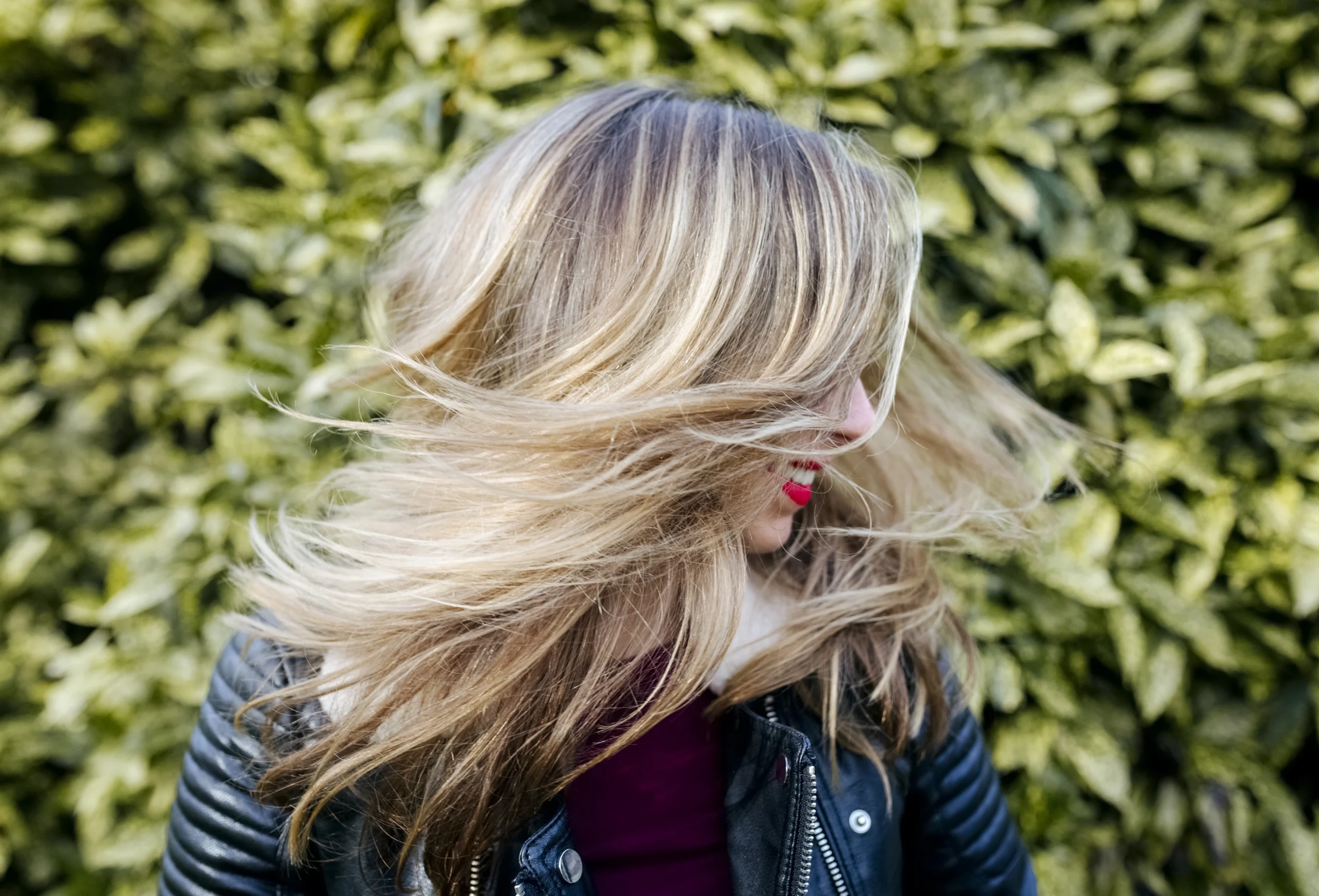Person with long, blonde hair wearing a black leather jacket, turning their head, with blurred motion against a leafy green background. - Trios Aveda Salon | Fort Collins, CO