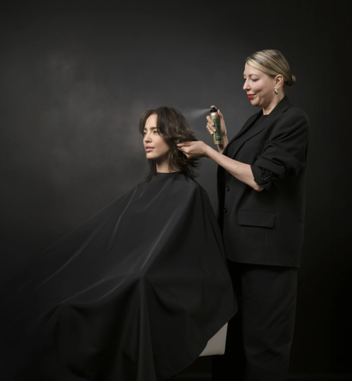 A hairstylist in black clothes sprays hair product on a seated woman with shoulder-length brown hair against a dark background. - Trios Aveda Salon | Fort Collins, CO