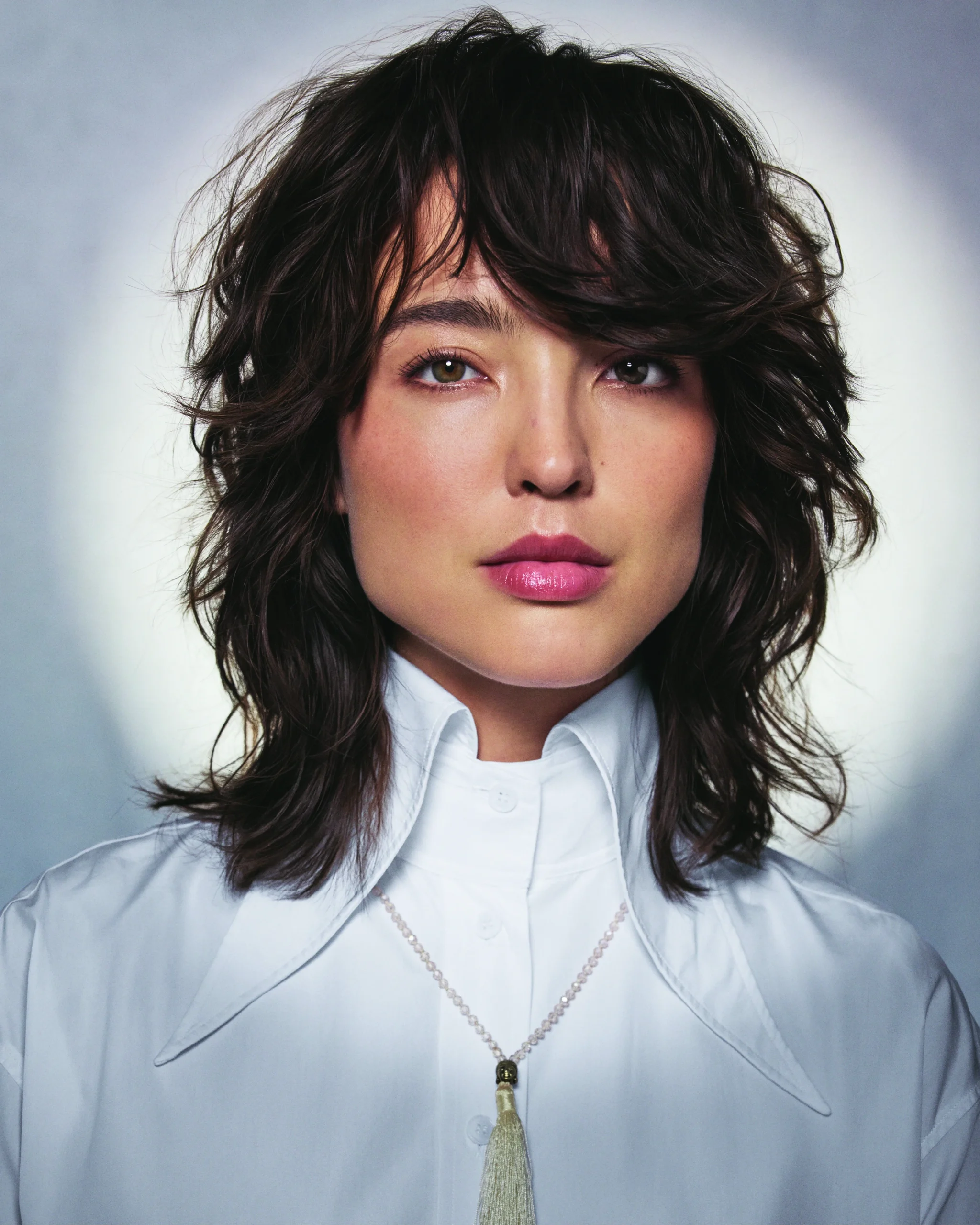 Person with wavy, medium-length dark hair wearing a white collared shirt and a necklace with a tassel, photographed against a pale background. - Trios Aveda Salon | Fort Collins, CO