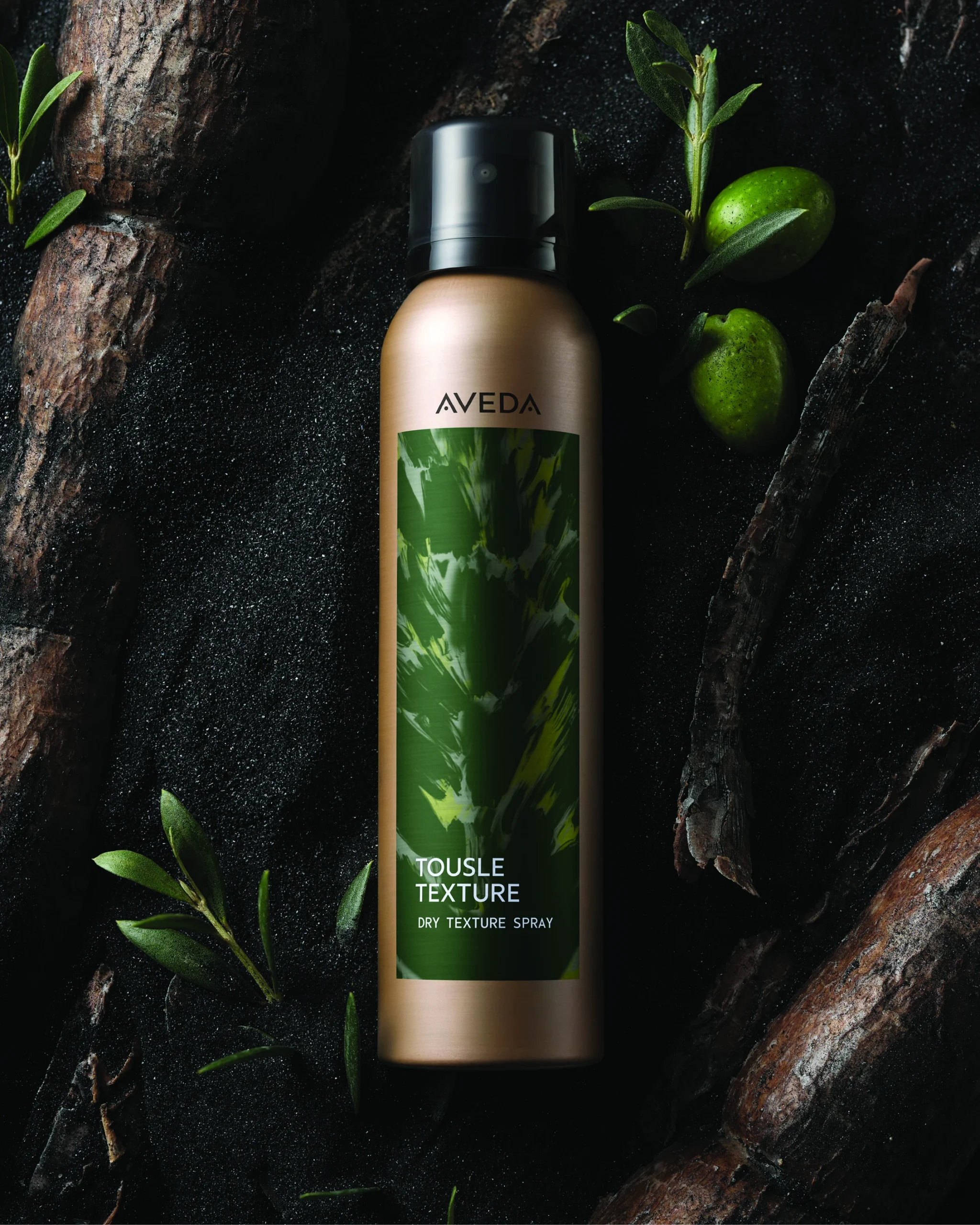 Aveda Tousle Texture Dry Texture Spray can placed on a dark surface with green leaves and two green fruits nearby. - Trios Aveda Salon | Fort Collins, CO