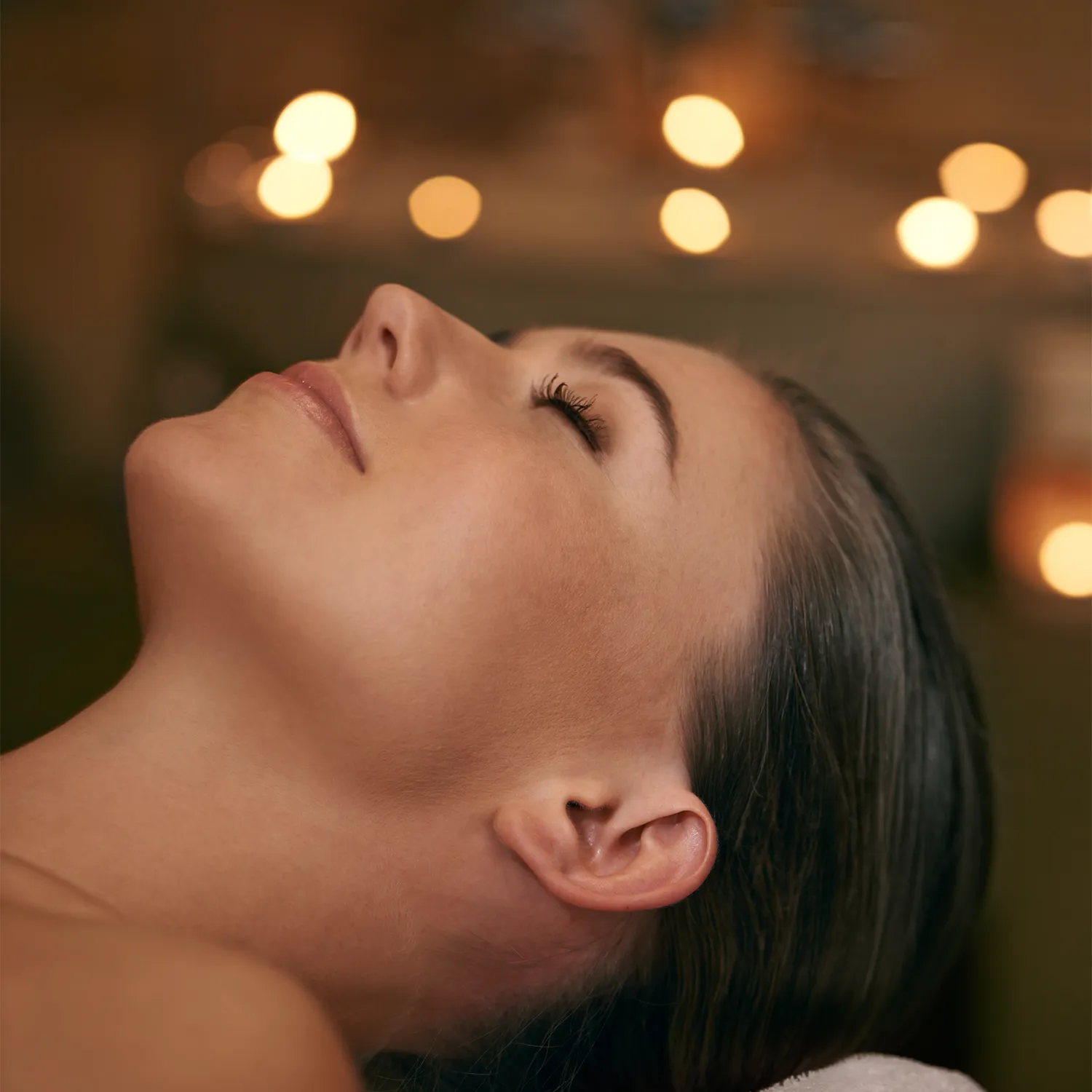 A woman lies on her back with eyes closed, head tilted upward, in a dimly lit room with blurred warm lights in the background. - Trios Aveda Salon | Fort Collins, CO